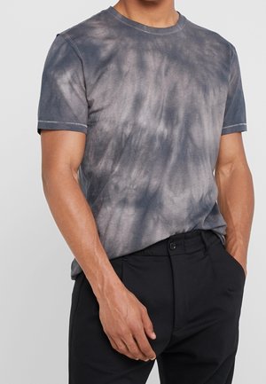 Hombre con camiseta de manga corta gris y negra con diseño tie-dye y pantalones negros, con una mano en el bolsillo sobre un fondo liso.