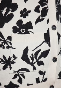 Cardigan in maglia nera a fiori con sfondo crema, tasca a costine e bottoni neri. Il motivo include varie design floreali.