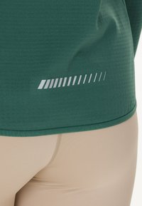 Groene sporttop met een gestructureerde afwerking, voorzien van een witte diagonale streepjespatroon nabij de zoom. Gecombineerd met beige leggings.