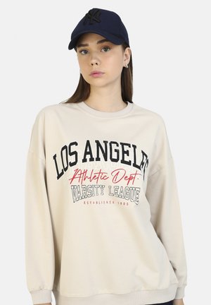 Beige Sweatshirt mit dem Druck "LOS ANGELES ATHLETIC DEPT. VARSITY LEAGUE" in Schwarz und Rot. Kombiniert mit einer marineblauen Baseballmütze mit einem Logo.