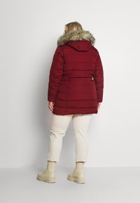 Bordeaux puffjacka med fuskpäls på huvan, horisontell quiltad design och två sidofickor. Bärs över krämfärgade byxor och beige stövlar.