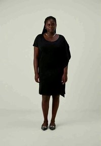 Crna haljina opuštenog kroja s kratkim rukavima, koja ima okrugli izrez i asimetričnu donju ivicu. Glatka tekstura; uparena s ravnim, dekorativnim cipelama.