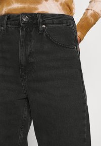 Černé džínové kalhoty s vysokým pasem, s knoflíkovým uzávěrem, bočními kapsami a klasickým designem pěti kapes. Texturovaná tkanina s jemným leskem.