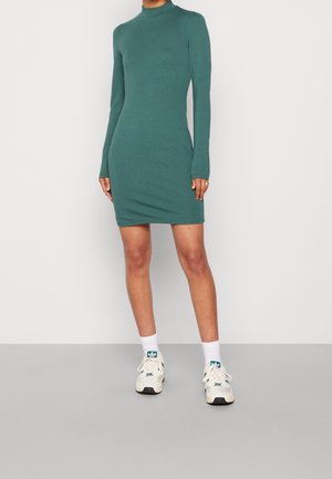 Woman wearing a fitted, long-sleeve green dress with a high neckline, white crew socks, and white Adidas sneakers, standing against a plain background.