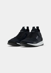 Paire de baskets de sport noires de la marque BOSS avec semelles blanches texturées exposées sur un fond gris clair uni.