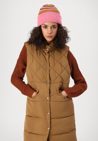 Gilet matelassé marron avec un col montant et des boutons pression, associé à un pull à manches en tricot rouille. Bonnet rayé rose et marron sur la tête.