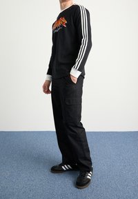 Svart långärmad tröja med vita detaljer och grafisk design, matchad med svarta cargobyxor och svarta sneakers med vita ränder.