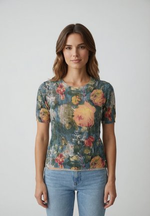 Donna con capelli castano ondulati che indossa una maglietta a maniche corte con motivo floreale e jeans azzurri, in piedi contro uno sfondo chiaro e uniforme.