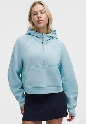 Blondine draagt een lichtblauwe capuchonsweater met voorrits en zak, gecombineerd met een zwarte rok.
