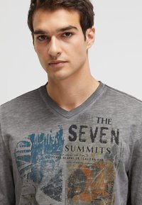 Graues V-Ausschnitt-T-Shirt mit buntem Grafikdruck und dem Text "DIE SIEBEN GIPFEL", strukturiertem Stoff und kontrastierendem Stich entlang des Ausschnitts.