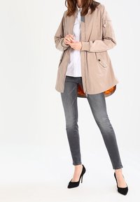 Chaqueta bomber ligera beige con cierre de cremallera, dos bolsillos laterales y forro gris. Combinada con jeans ajustados gris oscuro y tacones negros.