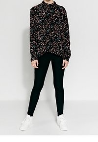 Svart blommig blus med rosa och blå mönster, har långa ärmar och en krage, tillsammans med svarta leggings och vita sneakers.