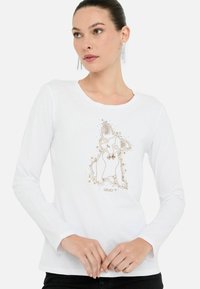 Tricou alb cu mânecă lungă, având un design grafic auriu al unui animal așezat, împodobit cu mici elemente decorative și text.