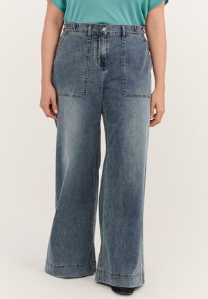 Weit geschnittene blaue Denim-Jeans mit vorderen aufgesetzten Taschen, getragen von einer Person in einem türkisfarbenen Oberteil und schwarzen Schuhen, stehend vor schlichtem Hintergrund.