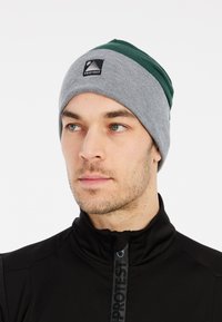Gorra de punto gris con acentos verdes, que presenta un pequeño parche de logo negro. El diseño es ajustado, mostrando una textura acanalada.