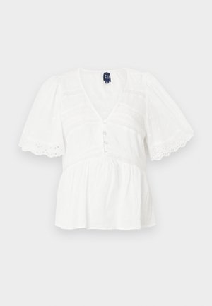 Blouse blanche avec des manches courtes bouffantes, décolleté en V, fermeture à boutons et détails texturés. Comprend des accents en dentelle et un design à ourlet évasé.