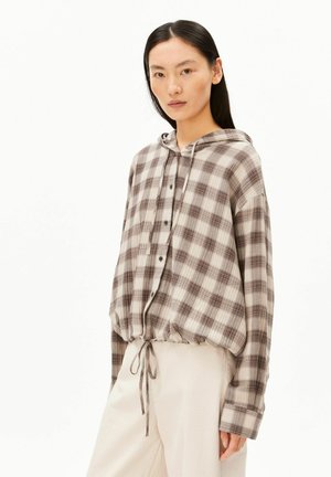 Vrouw draagt een losse bruine en beige geruite capuchonblouse met knopen en een koord in de taille, gecombineerd met brede beige broek, staand tegen een witte achtergrond.