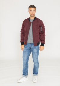 Bordowa kurtka bomberka z czarnymi ściągaczami, kieszenią na suwak na rękawie, zestawiona z szarym T-shirtem i niebieskimi dżinsami. Białe trampki.