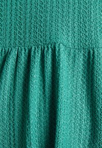 Tecido texturizado em verde-água com padrão zigzag e detalhes franzidos na cintura. O material parece leve e ligeiramente iridescente.