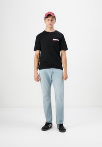 Nuori mies, jolla on päässään punainen hattu, musta Jack & Jones -t-paita, vaaleansiniset farkut ja mustat lenkkarit, seisoo tasaisella valkoisella taustalla.