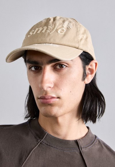Khaki baseball cap with embroidered "smile" text. Soft fabric, curved brim, and slight distressed accents. Model wearing a brown sweatshirt.