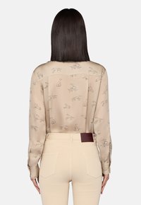 Donna con capelli scuri e lisci indossa pantaloni beige e una camicia a maniche lunghe di un beige chiaro, decorata con piccole illustrazioni di cani, rivolta di spalle.