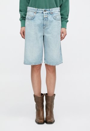 Hellblaue Denim-Bermudashorts mit geradem Schnitt, fünf Taschen und sichtbaren Nähten. Kombiniert mit braunen Lederknöchelstiefeln.