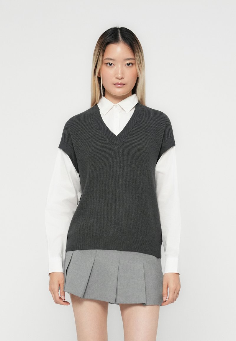 Gilet grigio lavorato a maglia con scollatura a V, indossato sopra una camicia bianca con colletto; abbinato a una gonna grigia plissettata. Design minimal con linee pulite.