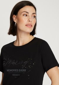 Czarna koszulka z krótkim rękawem z graficznym nadrukiem w srebrnym i szarym kolorze, z napisem "Tworzenie Wspomnień Każdego Weekendu", ozdobiona małymi metalowymi akcentami.