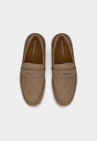 Ζευγάρι ανοιχτόχρωμες σουέντ μοκασίνια με λευκές σόλες, με την επωνυμία "KARL LAGERFELD" στις λουρίδες και στις εσωτερικές σόλες, απεικονισμένο από ψηλά σε λευκό φόντο.