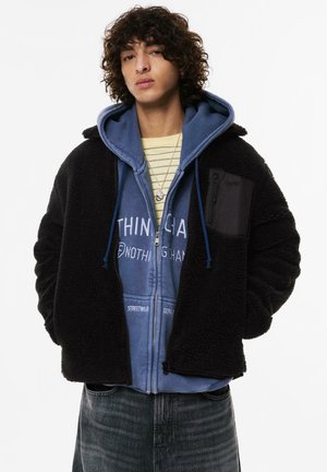 Veste en polaire noire à surface texturée, superposée sur un sweat à capuche zippé bleu avec des lettres blanches, complétée par une chemise rayée en dessous.