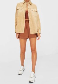 Beige corduroy overhemdjack met twee borstzakken, gedragen over een bruine cropped tanktop en bruine shorts, gecombineerd met witte sneakers.