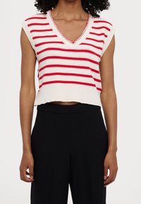Kobieta w białym bezrękawnym swetrze z czerwonymi poziomymi paskami i dekoltem w serek, zestawionym z czarnymi spodniami o wysokim stanie.