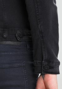 Detail paže a pasu osoby oblečené v černé džínové bundě a tmavě modrých džínách, na které jsou vidět knoflíky bundy a tetování na zápěstí.