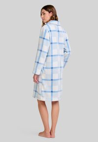 Mujer de pie descalza, vistiendo una bata de felpa suave hasta la rodilla con un patrón de cuadros azul y blanco, de espaldas contra un fondo gris.