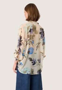 Blusa fluida e trasparente color beige chiaro con motivo floreale ad acquerello nei colori blu, verde e marrone; presenta maniche larghe con lacci.
