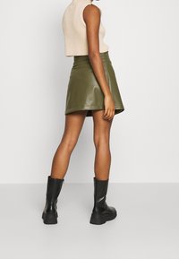 Jupe mini en simili cuir vert olive avec une forme évasée, assortie à un haut sans manches beige à côtes et des bottes à semelle épaisse noires.