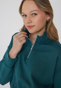 Zielona bluzka z zamkiem na suwak o wysokim kołnierzu, miękka w dotyku, z prążkowanymi mankietami. Model trzyma zamek, prezentując zbliżenie wzoru.