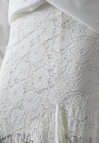 Hvid blondekjole med blomster mønstre, struktureret med indviklede detaljer. Stoffet har en scallop kant ved kanten.