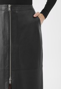 Zwarte leren rok met een zilveren rits aan de voorkant en een hand in de rechterzak, gedragen met een zwart langmouwen top op een witte achtergrond.