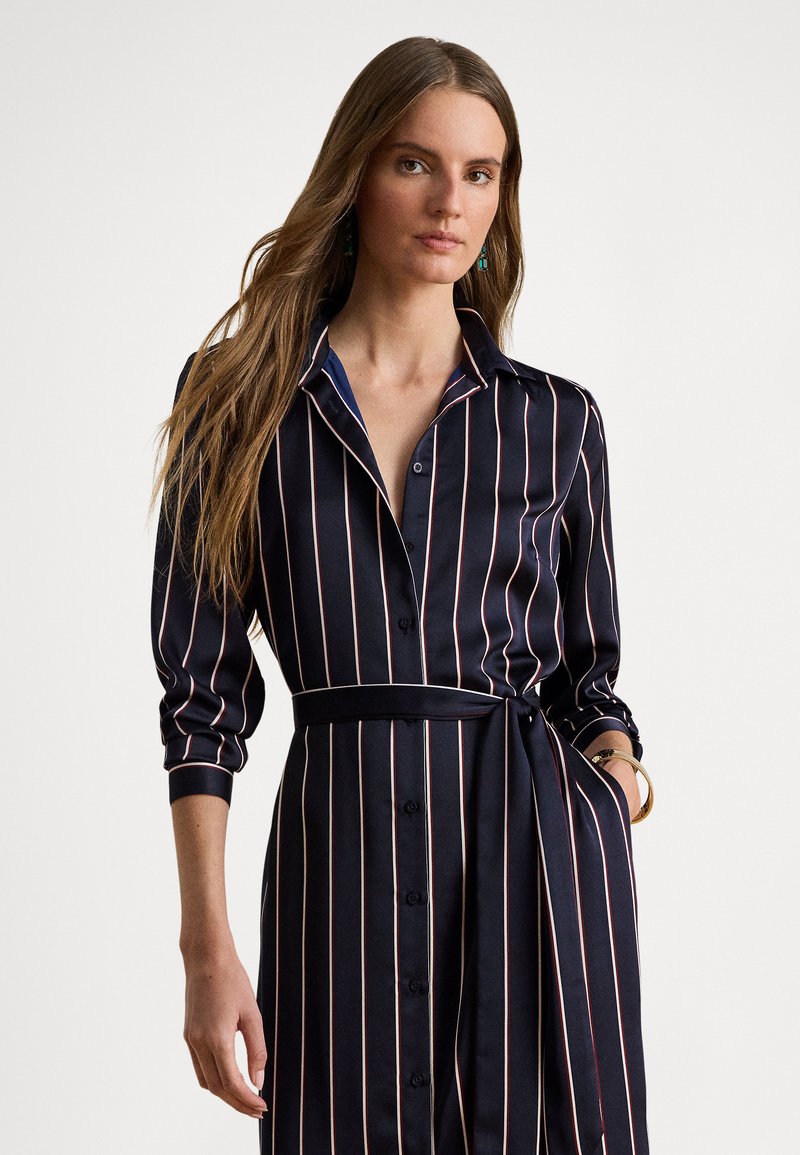 Φόρεμα πουκάμισο σε ναυτικό μπλε με κάθετες ροζ ρίγες, κουμπωτό μπροστά, με ορισμένο γιακά και με ζώνη στη μέση, με μανίκια τριών τετάρτων.