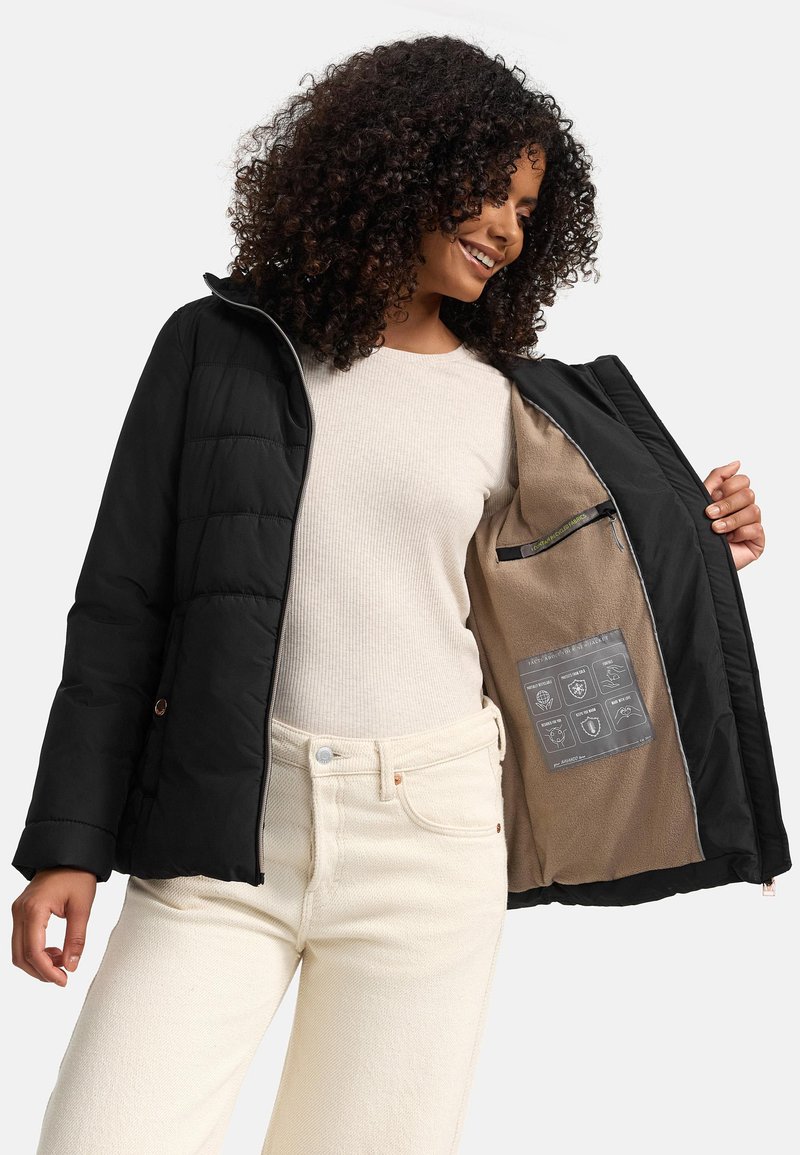 Black puffer jacket with a zip front, featuring a beige lined interior with care instructions. Ribbed cream top and light-colored pants.