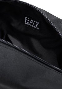 Pouch sintético negro con un exterior estampado y cierre de cremallera. El interior revela una etiqueta de la marca: "EA7 Emporio Armani."