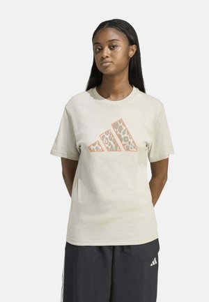 Mujer con cabello largo y oscuro que lleva una camiseta beige con un logo de estampado de leopardo y pantalones cortos oscuros con un pequeño logo blanco.