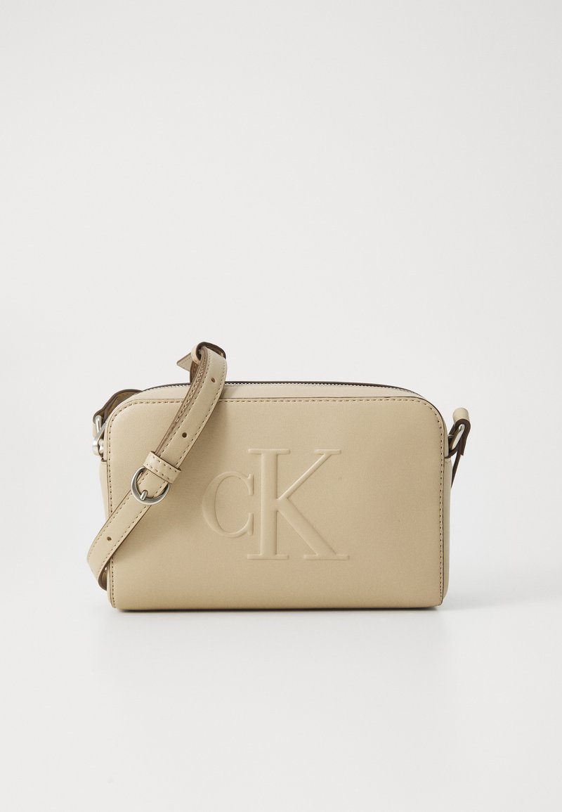 Beige skinnveske med overdimensjonert "cK"-logo. Har glidelås, avtakbar stropp og en jevn tekstur. Kompakt rektangulær form.
