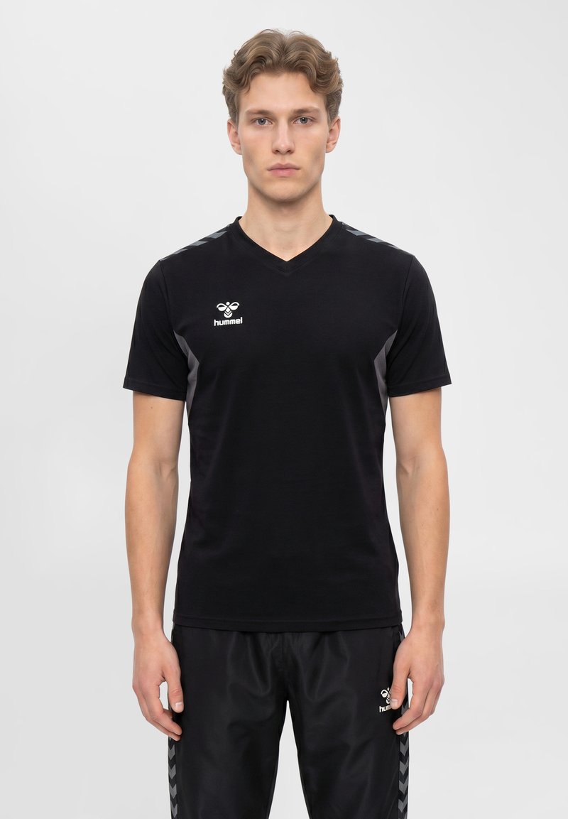 Junger Mann mit schwarzem hummel Sport-T-Shirt und schwarzen Hosen steht vor einem schlichten hellen Hintergrund.
