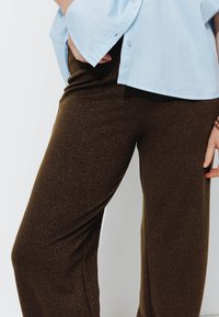 Pantalons marron amples en tissu doux et texturé, avec de subtils paillettes. Associés à une chemise à manches courtes et à boutons de couleur bleu clair.