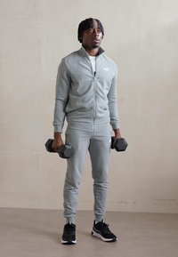 Survêtement de sport zippé gris, baskets noires, tenant des haltères hexagonaux. Tissu lisse, design ajusté, logo visible sur la poitrine.