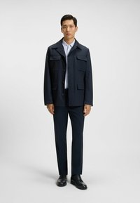 Miehen tummansininen takki, jossa on neljä etutaskua, nappisuljenta ja kärjellinen kaulus, yhdistettynä samansävyisiin tummiin housuihin ja mustiin kenkiin.