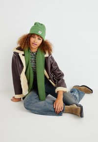 Veste en shearling marron, bonnet et écharpe en tricot verts, chemise rayée, jeans bleus et bottines beiges. Modèle assis contre un fond blanc.